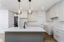 Modern kitchen featuring a central island with an undermount sink and matte black faucet, light grey flat-panel cabinetry, white countertops, and a white tile backsplash - 