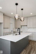 Kitchen featuring a large central island with a white countertop and dark gray base, under-mount double basin sink with a matte black gooseneck faucet, light gray flat-panel cabinetry, white subway tile backsplash, and wood-finish flooring - 