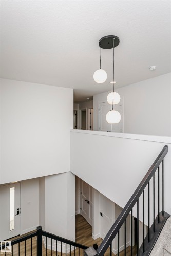 Modern tiered pendant lighting illuminates the open interior featuring a black metal and wood-finish staircase railing, white panel doors with black hardware, and wood-finish flooring - 6210 Dansereau Crescent, Beaumont, AB 