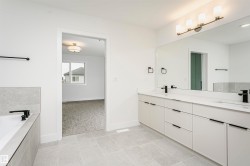 Spacious bathroom featuring light gray floor tiling, a deep soaking tub with gray tile surround, a dual vanity with white countertops and matte black fixtures, a large mirror, and a contemporary four-light fixture - 