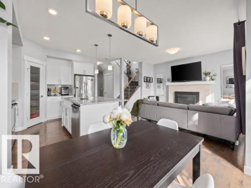 Open-concept interior featuring wood-finish flooring throughout, a kitchen island with granite countertops, stainless steel appliances, a built-in fireplace with a stone surround, and an integrated staircase with dark wood-finish treads - 607 40 Street, Edmonton, AB 