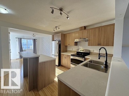 Kitchen featuring wood-finish cabinetry, light countertops, stainless steel appliances, a double basin sink with a black coiled faucet, and a breakfast bar - #13 3751 12 Street, Edmonton, AB 