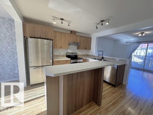 Kitchen featuring wood-finish cabinetry, stainless steel appliances, light-toned countertops, a breakfast bar, and wood-finish flooring - #13 3751 12 Street, Edmonton, AB 