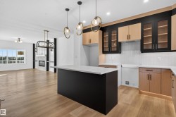 Open-concept kitchen and living area featuring wood-finish flooring, a kitchen island with a light-toned countertop, two-toned cabinetry with glass-front uppers, a white subway tile backsplash, and a modern fireplace with a marble-look surround - 