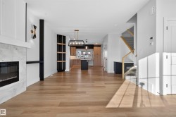 Open-concept living space featuring wood-finish flooring, a modern fireplace with a light-toned surround, a contemporary chandelier, a staircase with wood railings, and built-in shelving - 