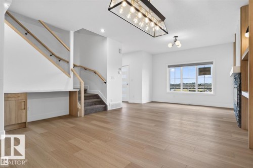 Spacious living area featuring light wood-finish flooring, a contemporary linear chandelier, and a large multi-pane window - 41 River Birch Way, St. Albert, AB 