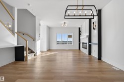 Open-concept living space featuring wood-finish flooring, a contemporary fireplace with tile surround, a modern black and gold chandelier, a staircase with light wood railings, and a large window providing ample natural light - 