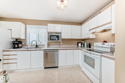 L-shaped kitchen featuring white cabinetry, light-toned countertops, tile flooring, a stainless steel dishwasher, and a white electric range - 11831 9 Avenue, Edmonton, AB 