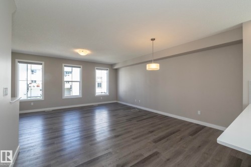 Open-concept living space featuring wood-finish flooring, three large windows, a contemporary pendant light fixture, a semi-flush mount ceiling light, and neutral wall tones - 2610 Maple Way, Edmonton, AB 