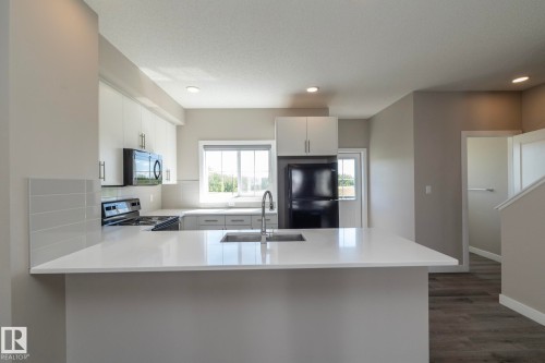 Kitchen featuring an expansive white countertop island with an undermount sink, white upper and lower cabinetry, black appliances, and light wood-finish flooring - 2610 Maple Way, Edmonton, AB 