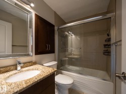 Bathroom featuring a granite-finish countertop with an undermount sink, a framed mirror with overhead lighting, a dark wood-finish wall cabinet, a white toilet, and a bathtub with a sliding glass enclosure - 