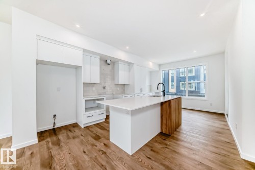 Open-concept kitchen featuring white shaker cabinetry, an expansive center island with a wood-finish accent panel, quartz countertops, a matte black gooseneck faucet, and a large window - #67 5 Rondeau Dr, St. Albert, AB - Indoor Photo Showing Kitchen