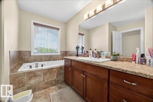 Bathroom featuring a built-in jetted tub with tile surround, a double vanity with an integrated sink, granite-finish countertops, wood-finish cabinetry, and a large mirror with overhead lighting - 1411 30 Avenue, Edmonton, AB 