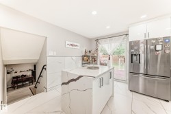 Contemporary kitchen featuring a central island with a waterfall countertop and integrated storage, stainless steel refrigerator, white gloss cabinetry, and large format porcelain tile flooring - 