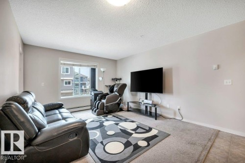 Living area featuring a large window, neutral wall tones, textured ceiling, and mixed flooring with carpet and tile - 219 1820 Rutherford Road, Edmonton, AB 