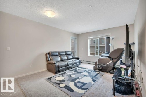 Bright living area featuring neutral wall tones, carpet flooring, a large window, and a flush mount ceiling light fixture - 219 1820 Rutherford Road, Edmonton, AB 