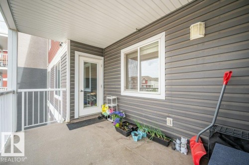 Covered concrete patio with grey horizontal siding, white trim windows, and a glass-paneled entry door - 219 1820 Rutherford Road, Edmonton, AB 