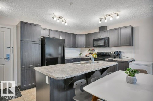 Kitchen featuring dark wood-finish cabinetry, black appliances, track lighting, and a kitchen island with a double basin sink - 219 1820 Rutherford Road, Edmonton, AB 