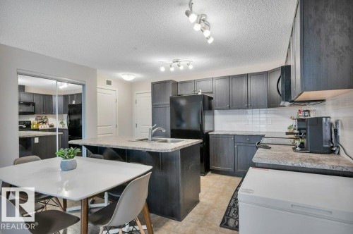 Modern kitchen featuring dark wood-finish cabinetry, granite-style countertops, an island with an undermount sink, and a white subway tile backsplash - 219 1820 Rutherford Road, Edmonton, AB 