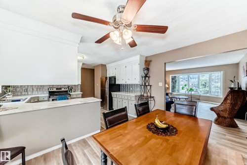 Open-concept dining area with wood-finish flooring, a ceiling fan with integrated lighting, and a pass-through to the kitchen featuring white cabinetry, stainless steel appliances, and a tiled backsplash - 9813 152 Street, Edmonton, AB 