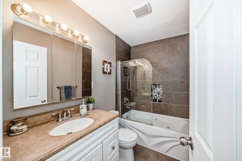 Bathroom featuring a vanity with a single basin sink and light-colored countertop, white cabinetry with silver hardware, a mirrored medicine cabinet with overhead lighting, a toilet, and a tiled shower-over-tub combination with a glass panel - 9813 152 Street, Edmonton, AB 