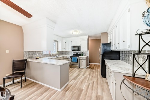 Open-concept kitchen featuring wood-finish flooring, white cabinetry, stainless steel appliances, marble-look countertops, and a decorative tile backsplash - 9813 152 Street, Edmonton, AB 