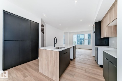 Contemporary kitchen featuring a central island with an integrated sink and matte black faucet, light wood-finish flooring, recessed lighting, expansive dark cabinetry, and light-toned upper cabinets - #66 5 Rondeau Dr, St. Albert, AB - Indoor Photo Showing Kitchen