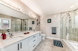 Bathroom featuring a dual vanity with white cabinetry and integrated sinks, a large wall-mounted mirror, a glass-enclosed shower, and light-toned tile flooring - 