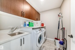 Dedicated utility space featuring stacked laundry appliances, an integrated sink, white base cabinetry, and wood-finish upper cabinets - 