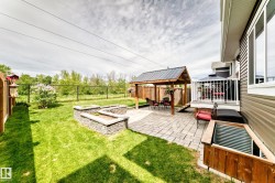 Expansive backyard featuring a stone paver patio, a covered wooden gazebo with a metal roof, two stone-clad raised garden beds, a second-story deck with white railing, and a window well with a wooden frame - 