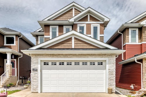 Contemporary exterior featuring brown siding, textured cedar shake accents, white trim, stone veneer detailing, and a prominent garage with paneling and window inserts - 3895 Robins Crescent, Edmonton, AB 