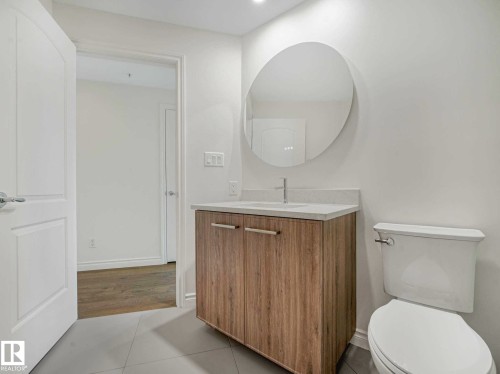 Contemporary vanity featuring a light wood-finish cabinet with sleek hardware, a white countertop, and a circular wall-mounted mirror - 902 10108 125 Street, Edmonton, AB 
