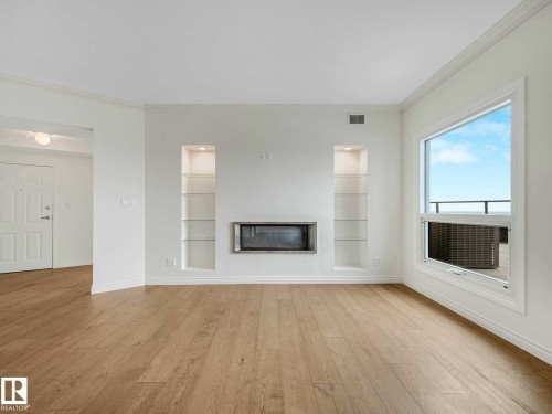 Spacious interior featuring wide plank wood-finish flooring, a contemporary fireplace, two illuminated display niches with glass shelving, a large window providing ample natural light, and crown molding - 902 10108 125 Street, Edmonton, AB 