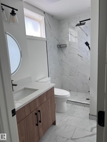 Bathroom featuring a wood-finish vanity with a white countertop and integrated sink, a rectangular shower with a clear glass enclosure, and white tile flooring - 11911 133 Avenue, Edmonton, AB 