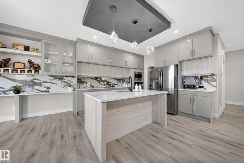 Contemporary kitchen featuring light wood-finish flooring, a central island with a white countertop, sleek gray cabinetry, a striking marbled backsplash, and recessed lighting with modern pendant fixtures - 449 Roberts Cres, Leduc, AB 