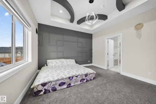 Bedroom featuring a tray ceiling with a contemporary pendant light fixture, a dark accent wall with geometric molding, light gray carpeting, and a large window with white trim - 449 Roberts Cres, Leduc, AB 