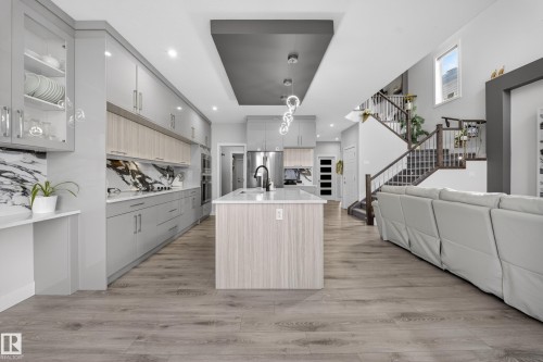 Open-concept kitchen featuring an oversized island, wood-finish flooring, a contemporary overhead light fixture, light gray cabinetry, and a dark marble-patterned backsplash - 449 Roberts Cres, Leduc, AB 