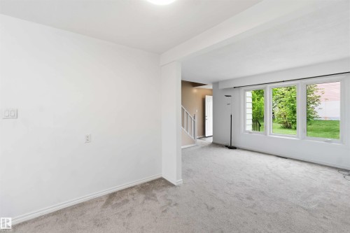 Spacious room featuring light gray carpeting, crisp white walls, multiple large windows, and a partially visible staircase with white railings - 17325 85 Avenue Nw, Edmonton, AB 