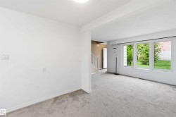 Spacious room featuring light gray carpeting, crisp white walls, multiple large windows, and a partially visible staircase with white railings - 