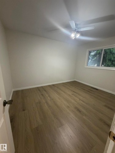 Spacious room featuring wood-finish flooring, neutral wall tones, a ceiling fan with integrated lighting, and a window with white trim - 9640 74 Avenue, Edmonton, AB 