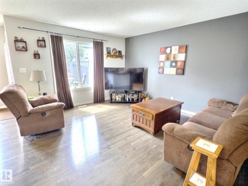 Spacious living area featuring wood-finish flooring, a large window with privacy curtains, and a contemporary accent wall - 126 Calvert Wynd, Fort Saskatchewan, AB 