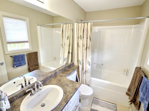 Bathroom featuring a single vanity with an oval undermount sink, a shower-over-tub combination with white acrylic surround, a toilet, and a window with blinds - 126 Calvert Wynd, Fort Saskatchewan, AB 
