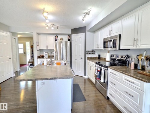 Kitchen featuring an island with an integrated sink, white cabinetry, stainless steel appliances, dark countertops, and wood-finish flooring - 126 Calvert Wynd, Fort Saskatchewan, AB 