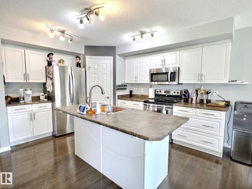 Bright kitchen featuring white cabinetry, dark wood-finish flooring, a stainless steel appliance suite, track lighting, and an island with an integrated sink - 126 Calvert Wynd, Fort Saskatchewan, AB 