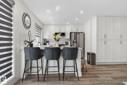 Contemporary kitchen featuring white shaker cabinetry, stainless steel appliances, a breakfast bar with stone countertop, subway tile backsplash, and wood-finish flooring - 16 9250 156 Avenue, Edmonton, AB 