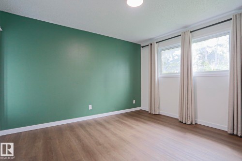 Interior room featuring a green accent wall, light wood-finish flooring, two large windows with drapes, white baseboards, and a flush-mount ceiling light - 10 Murray Crescent, St. Albert, AB 