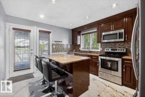 Kitchen featuring an island with stone countertop, stainless steel appliances, mosaic tile backsplash, wood cabinetry, and tiled flooring - 946 Jordan Crescent, Edmonton, AB 