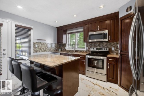 Kitchen featuring dark wood-finish cabinetry, stainless steel appliances, granite countertops, a mosaic tile backsplash, and a central island with bar seating - 946 Jordan Crescent, Edmonton, AB 