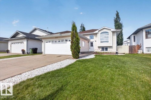 Exterior featuring light-colored siding, an attached two-car garage with white doors, a concrete driveway with white gravel accents, a front lawn, and mature trees - 946 Jordan Crescent, Edmonton, AB 