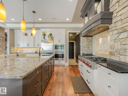 Gourmet kitchen featuring a stone accent wall, wood-finish flooring, a substantial kitchen island with a stone countertop, white cabinetry, and stainless steel appliances including a range and double oven - 2807 Wheaton Drive, Edmonton, AB 